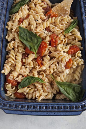 A blue ceramic baking dish filled with baked feta pasta, roasted grape tomatoes, and fresh basil leaves on an off-white background.