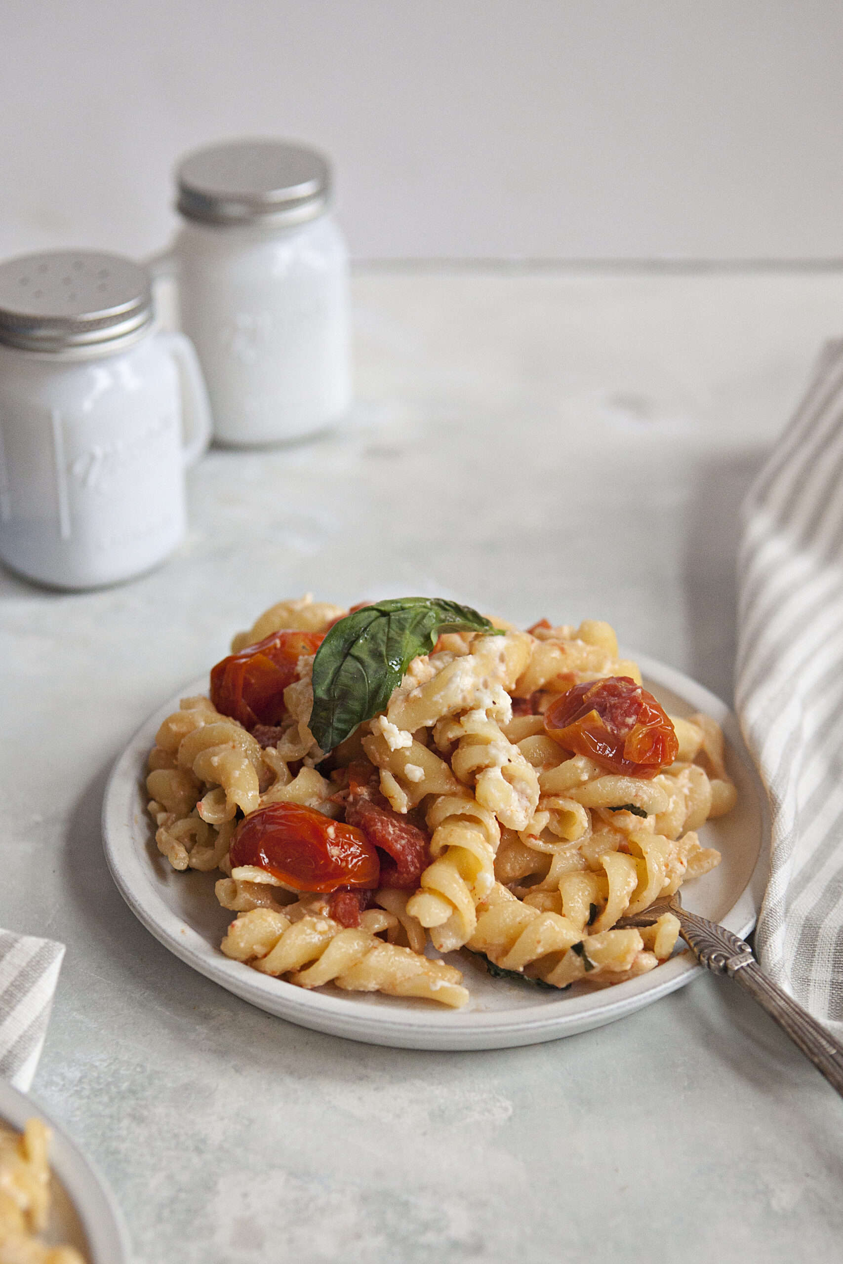 Baked feta pasta with roasted tomatoes and a whole basil leaf on a white plate next to a gray napkin and white salt and pepper shakers, on an off-white background.