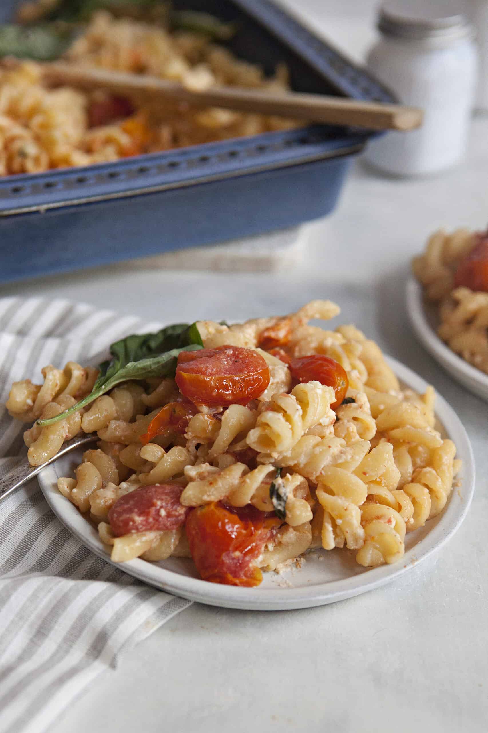 Baked feta pasta with roasted tomatoes and a whole basil leaf on a white plate next to a blue baking dish containing more feta pasta, near a gray napkin and a white salt shaker, on an off-white background.