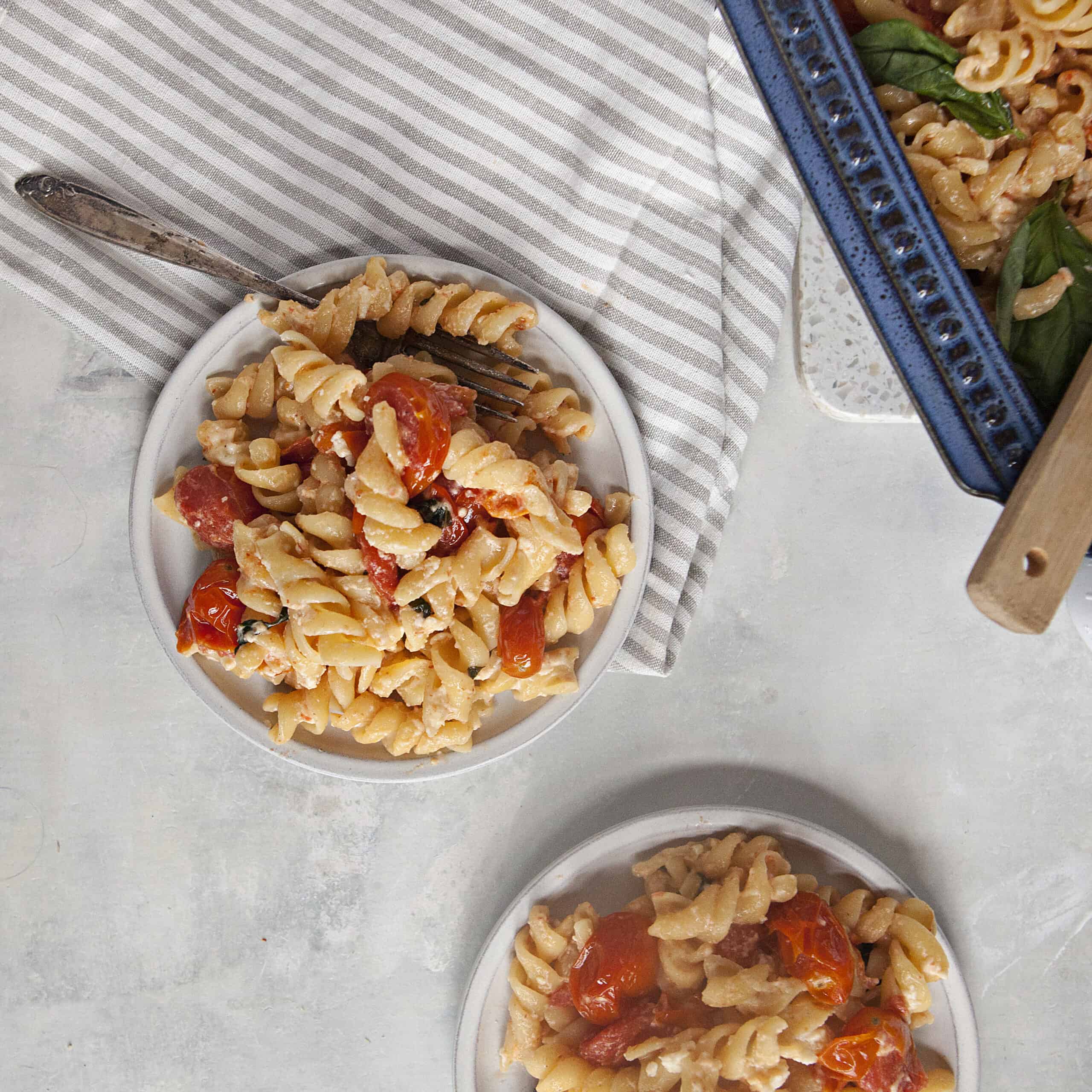 Two plates of baked feta pasta with roasted grape tomatoes next to a gray napkin and a blue baking dish with baked feta pasta on an off-white background.