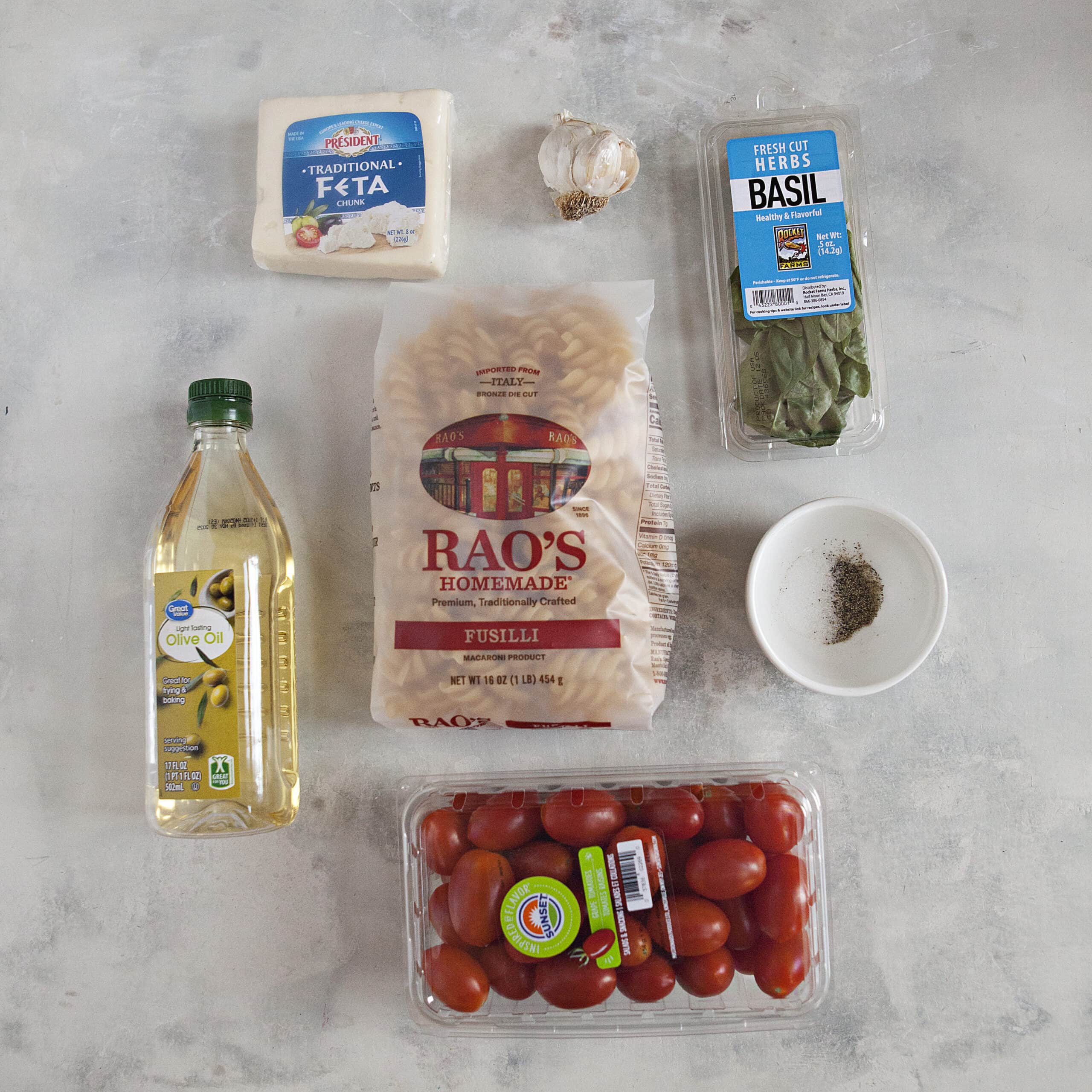 Ingredients for easy baked feta pasta with basil and garlic:  Fusilli pasta, olive oil, grape tomatoes, fresh basil, garlic, a block of feta cheese, salt, and pepper, on an off-white background.