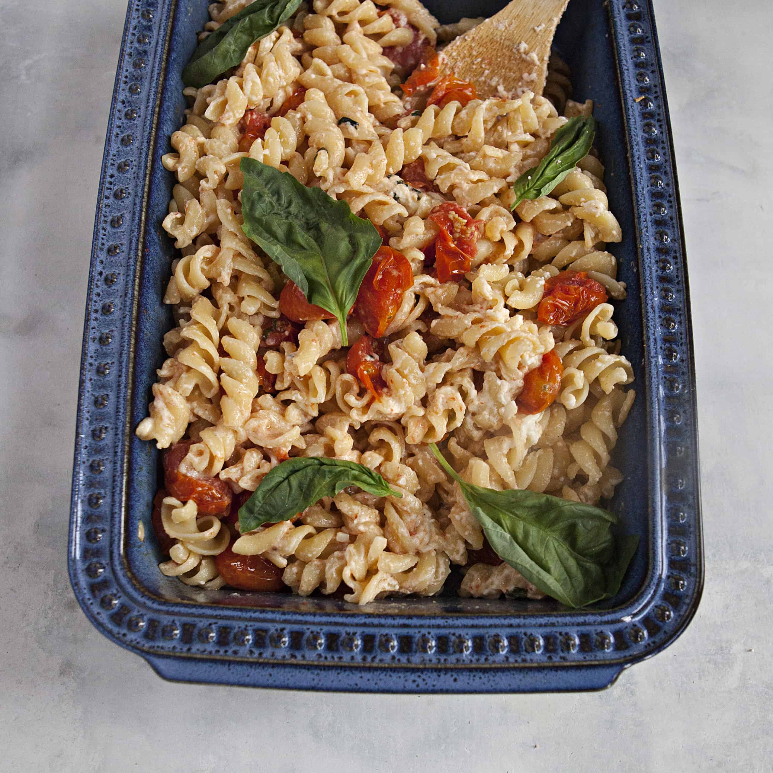 A blue ceramic baking dish filled with baked feta pasta, roasted grape tomatoes, and fresh basil leaves on an off-white background.