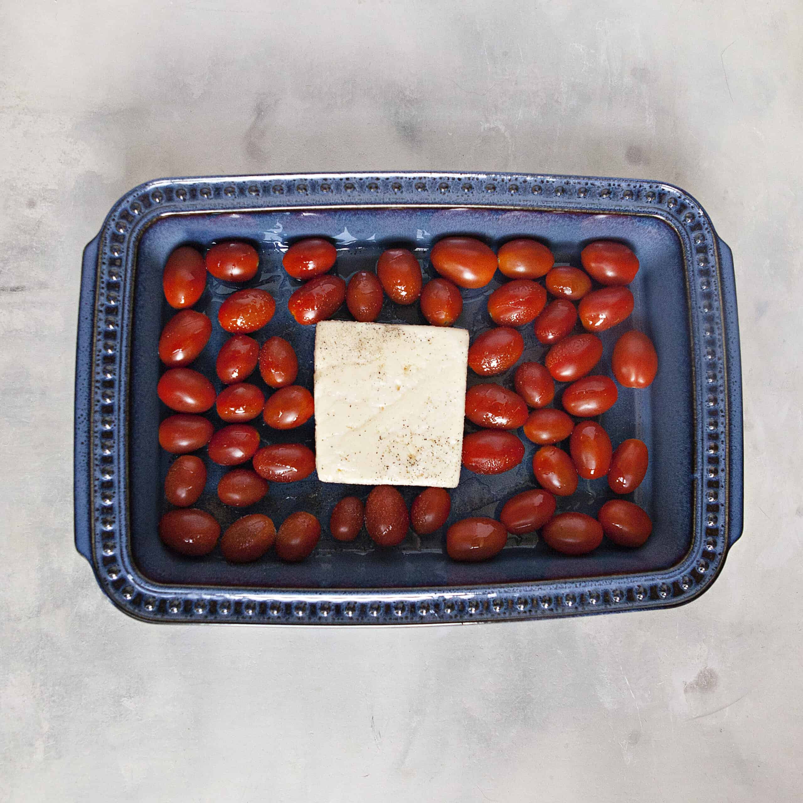 A blue ceramic baking dish containing a block of feta pasta, grape tomatoes, olive oil, salt and pepper on an off-white background.