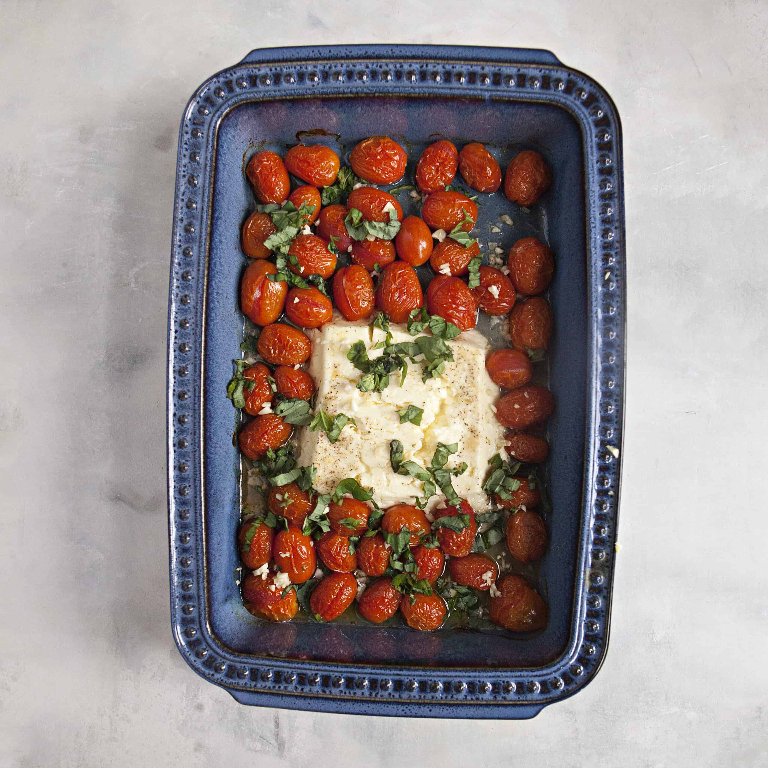 A blue baking dish containing a melted block of feta cheese, burst grape tomatoes, chiffonade of basil, and minced garlic on an off-white background.