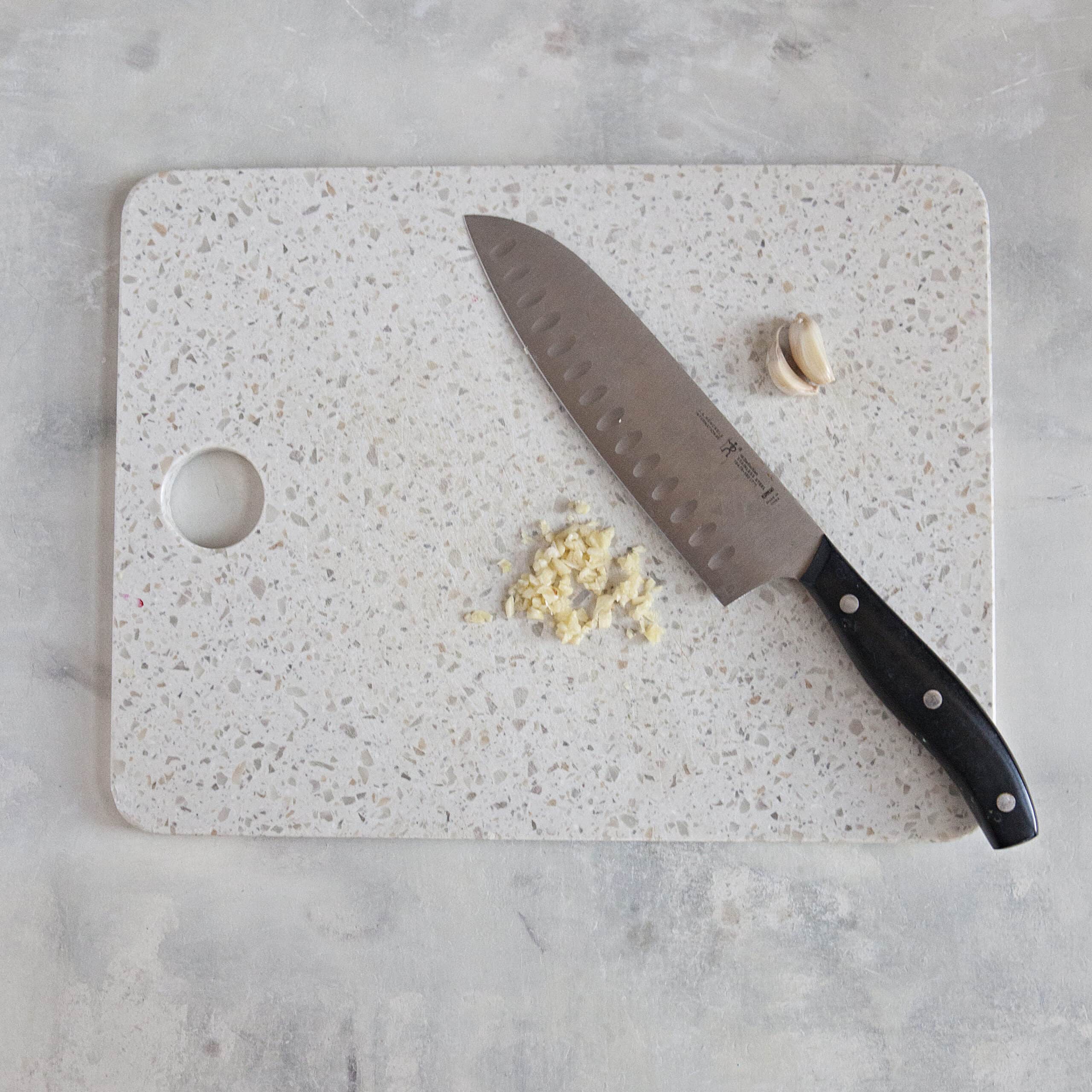 A white cutting board with minced garlic and cloves of garlic alongside a chef's knife on an off-white background.