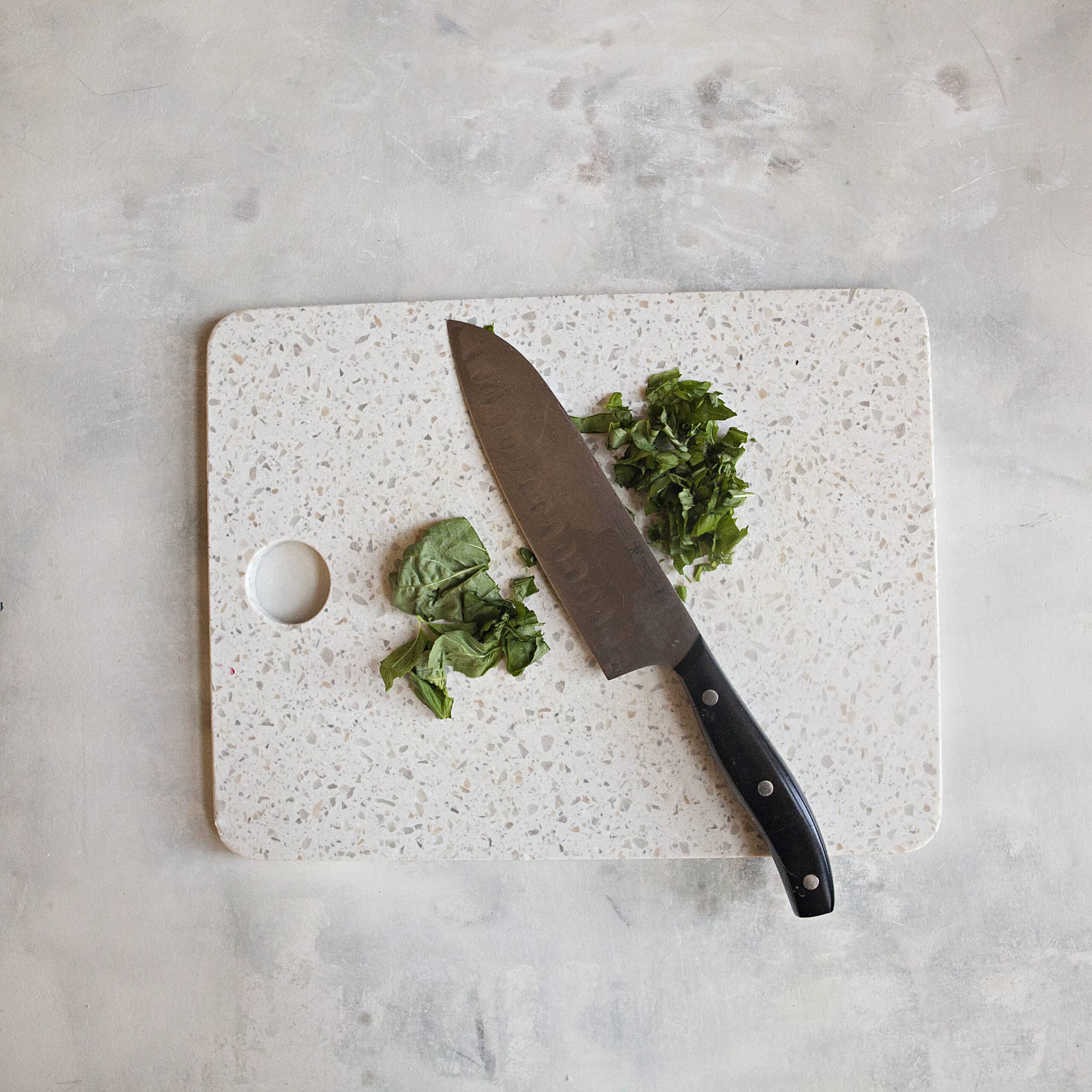 A white cutting board with chiffonade of basil and basil leaves alongside a chef's knife on an off-white background.