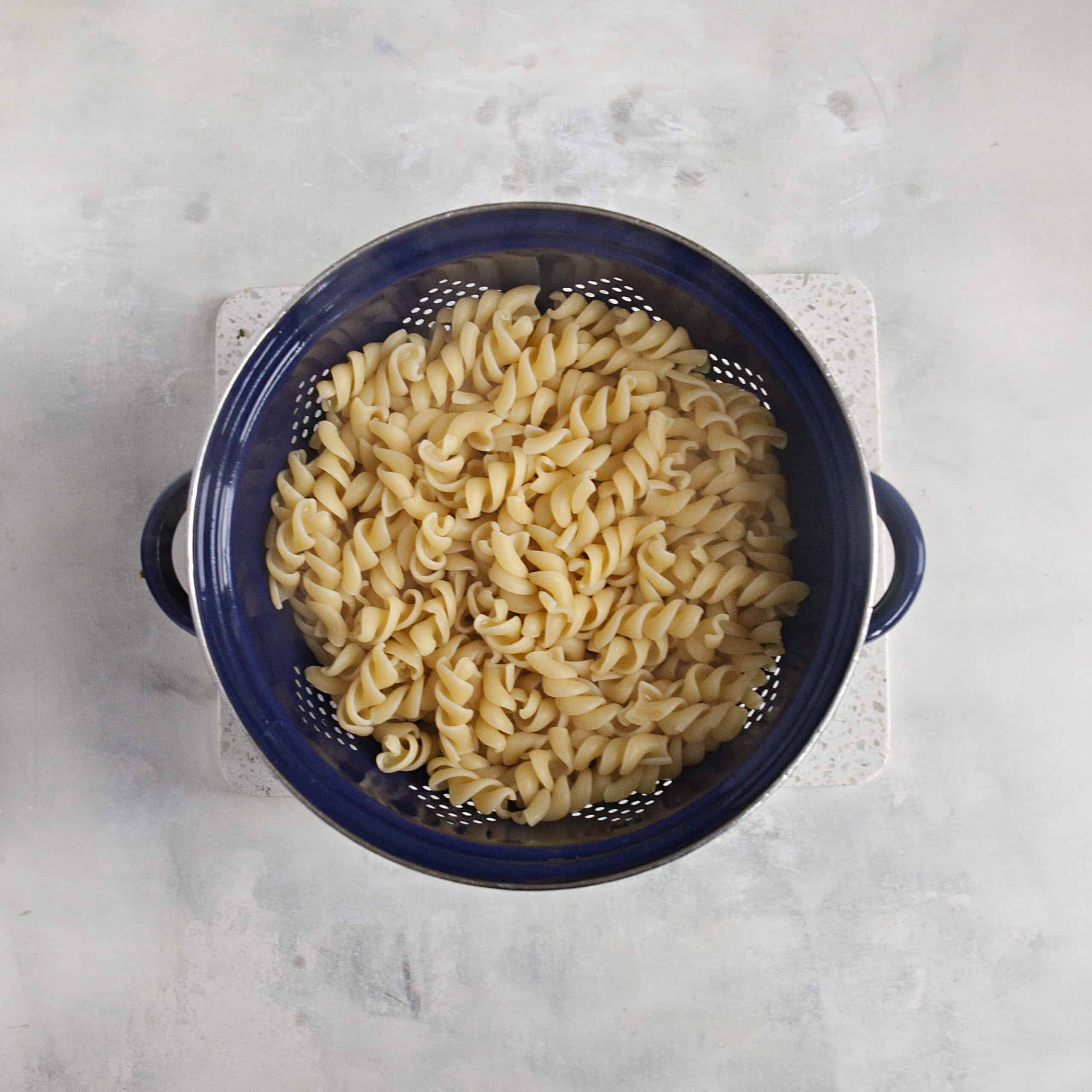 Cooked fusilli pasta in a navy blue colander on an off-white background.