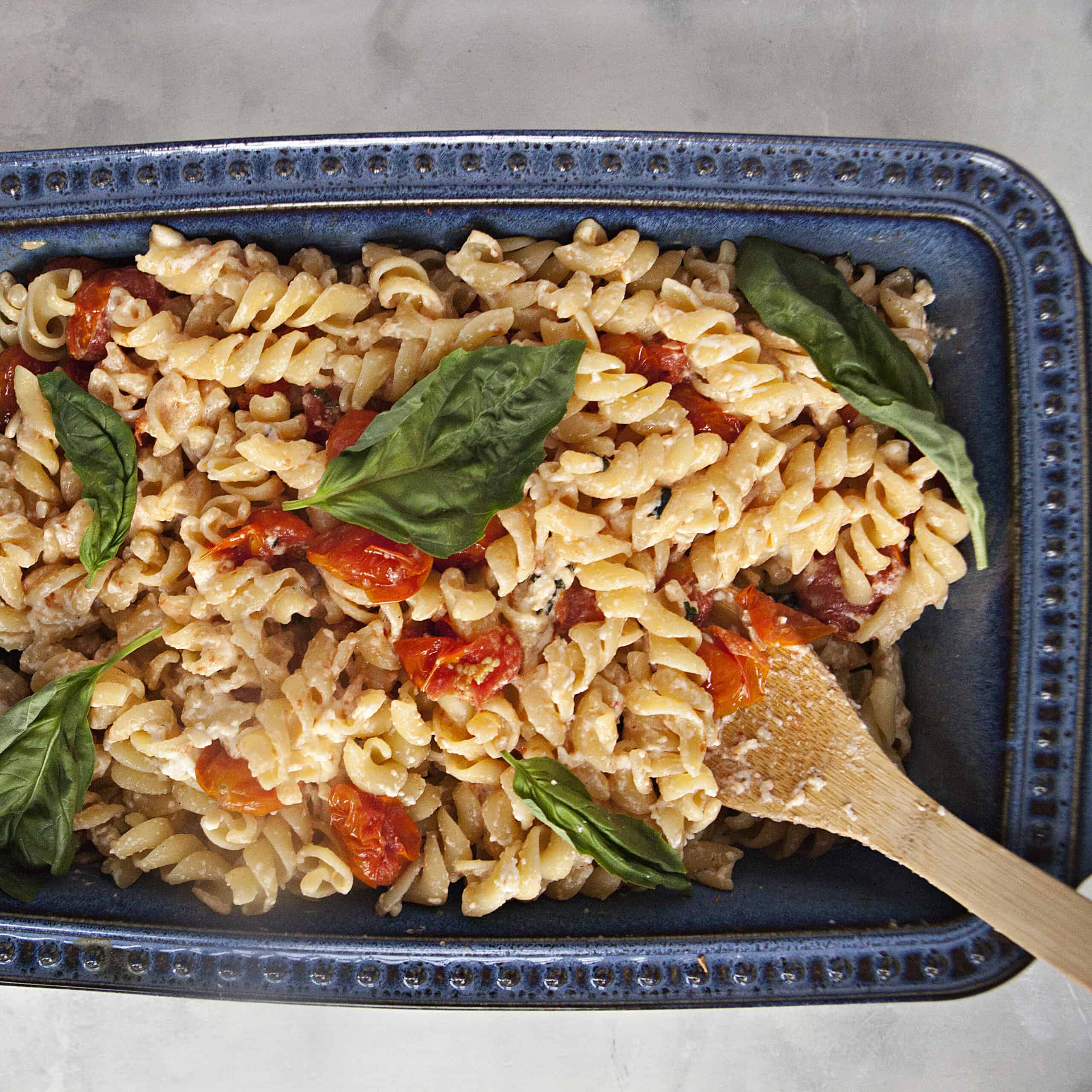 A blue ceramic baking dish filled with baked feta pasta, roasted grape tomatoes, and fresh basil leaves on an off-white background.