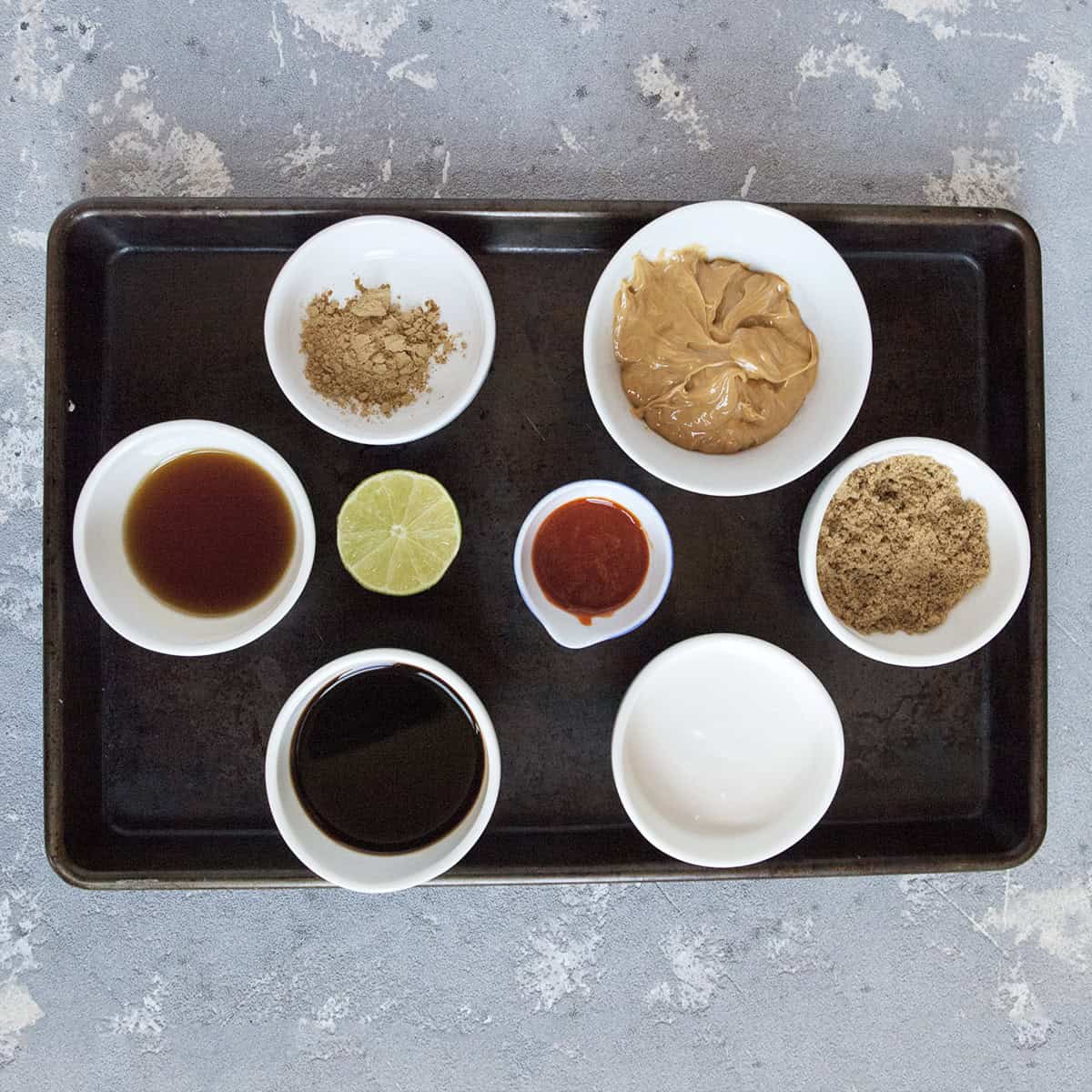 Ingredients for peanut dipping sauce:  Creamy peanut butter, soy sauce, ground ginger, brown sugar, fish sauce, coconut milk, sriracha, half a lime, in small white bowls on a dark brown baking sheet on a gray background.