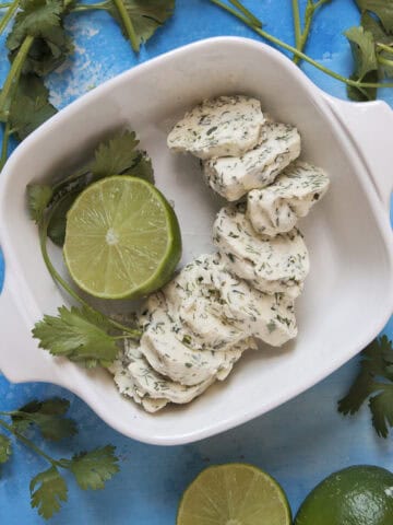 Round pieces of cilantro-lime herb compound butter in a baking dish with half a lime and a sprig of cilantro, on a blue background with more fresh cilantro and sliced limes.