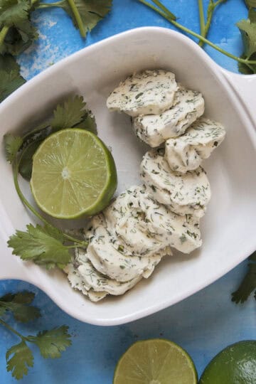 Round pieces of cilantro-lime herb compound butter in a baking dish with half a lime and a sprig of cilantro, on a blue background with more fresh cilantro and sliced limes.