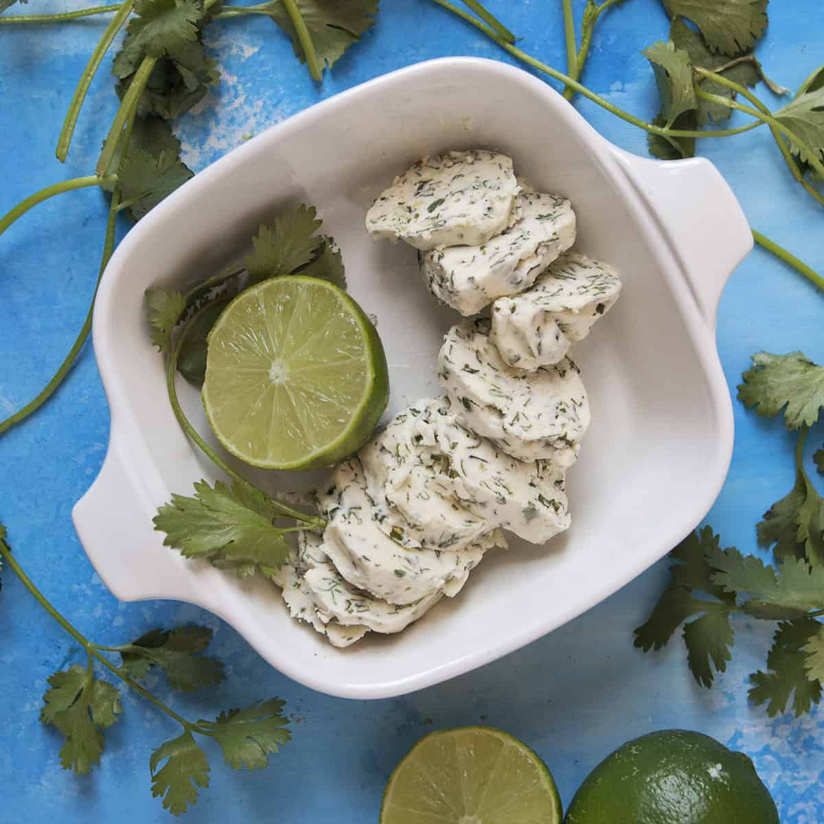 Round pieces of cilantro-lime herb compound butter in a baking dish with half a lime and a sprig of cilantro, on a blue background with more fresh cilantro and sliced limes.