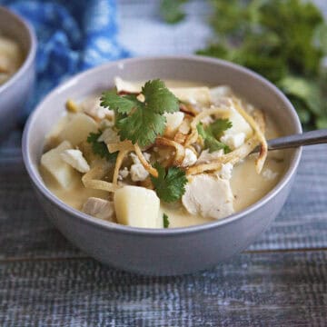 Cozy green chile chicken soup in a gray bowl with a spoon, topped with fried tortilla strips, crumbled Cotija cheese, and pieces of fresh cilantro, on a wooden table next to a blue napkin and more sprigs of cilantro.