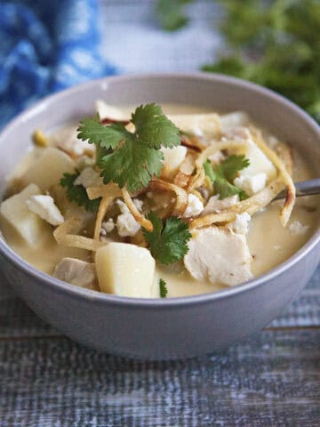 Cozy green chile chicken soup in a gray bowl with a spoon, topped with fried tortilla strips, crumbled Cotija cheese, and pieces of fresh cilantro, on a wooden table next to a blue napkin and more sprigs of cilantro.