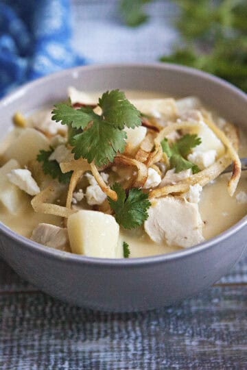 Cozy green chile chicken soup in a gray bowl with a spoon, topped with fried tortilla strips, crumbled Cotija cheese, and pieces of fresh cilantro, on a wooden table next to a blue napkin and more sprigs of cilantro.