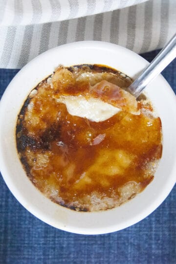 Chai-spiced creme brulee in a white ramekin with a caramelized sugar top, with a spoon, on a blue background with a gray napkin.