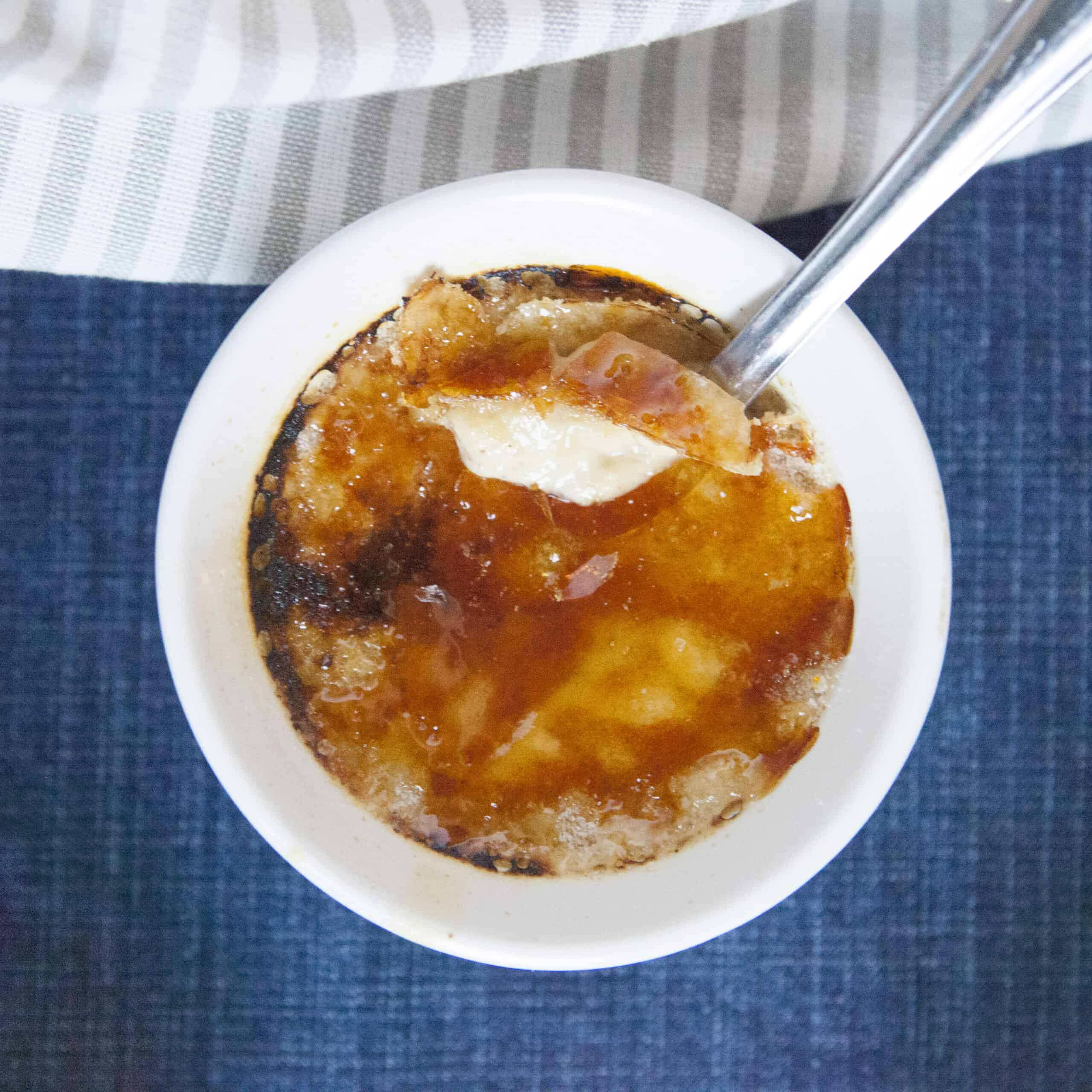 Chai-spiced creme brulee in a white ramekin with a spoon on a blue background with a gray napkin.