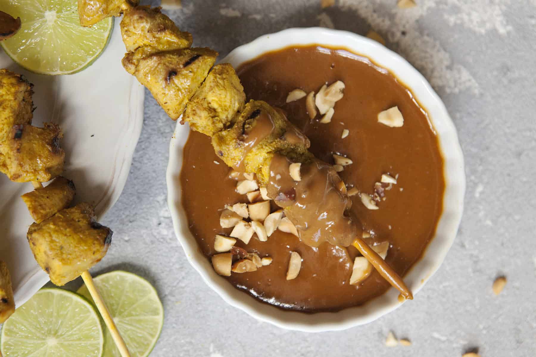 Thai-style grilled chicken satay skewers on a white scalloped plate alongside a bowl of peanut dipping sauce and chopped peanuts, slices of lime throughout the photo, on a gray background.