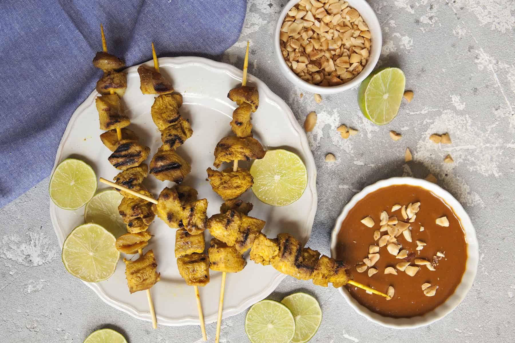 Thai-style grilled chicken satay skewers on a white scalloped plate alongside a bowl of peanut dipping sauce and chopped peanuts, slices of lime throughout the photo, on a gray background with a blue napkin.