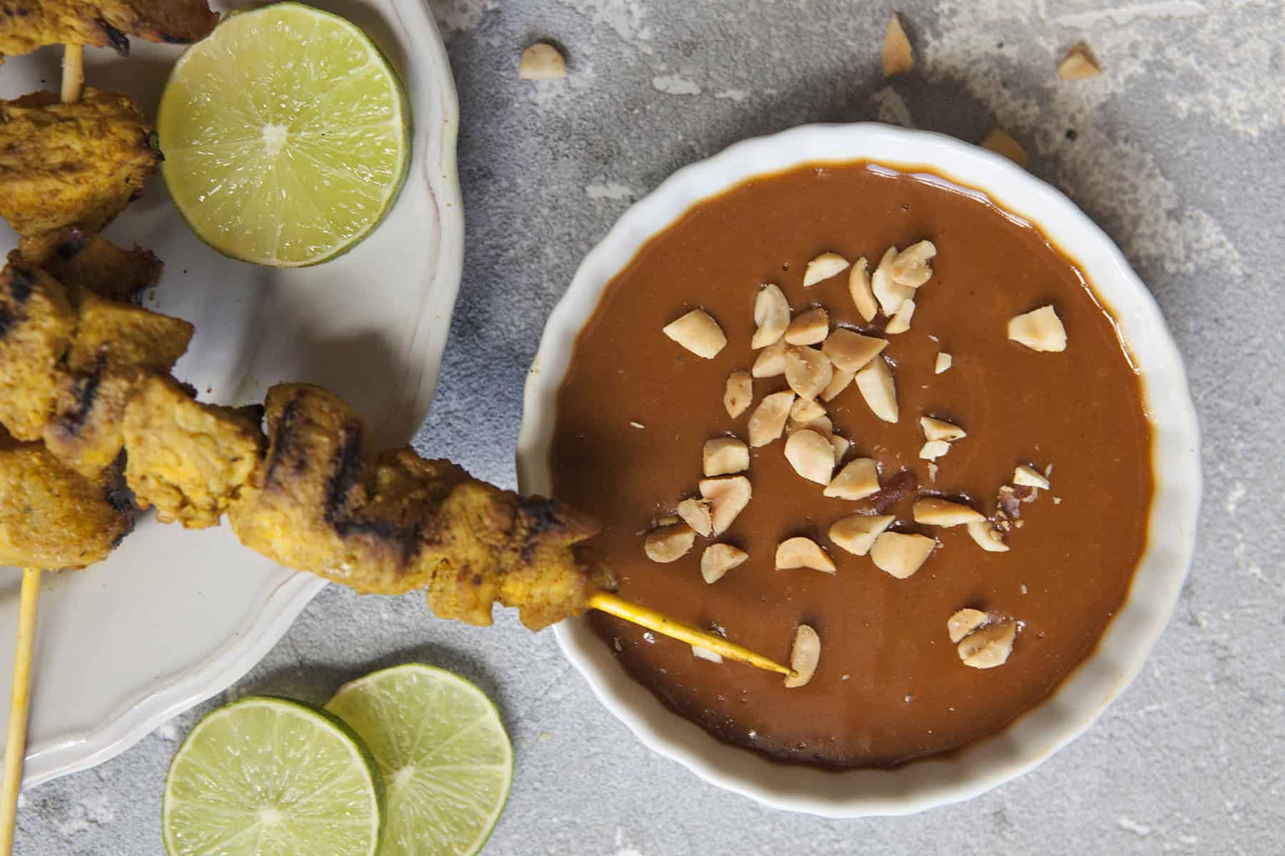 Thai-style grilled chicken satay skewers on a white scalloped plate alongside a bowl of peanut dipping sauce and chopped peanuts, slices of lime throughout the photo, on a gray background.