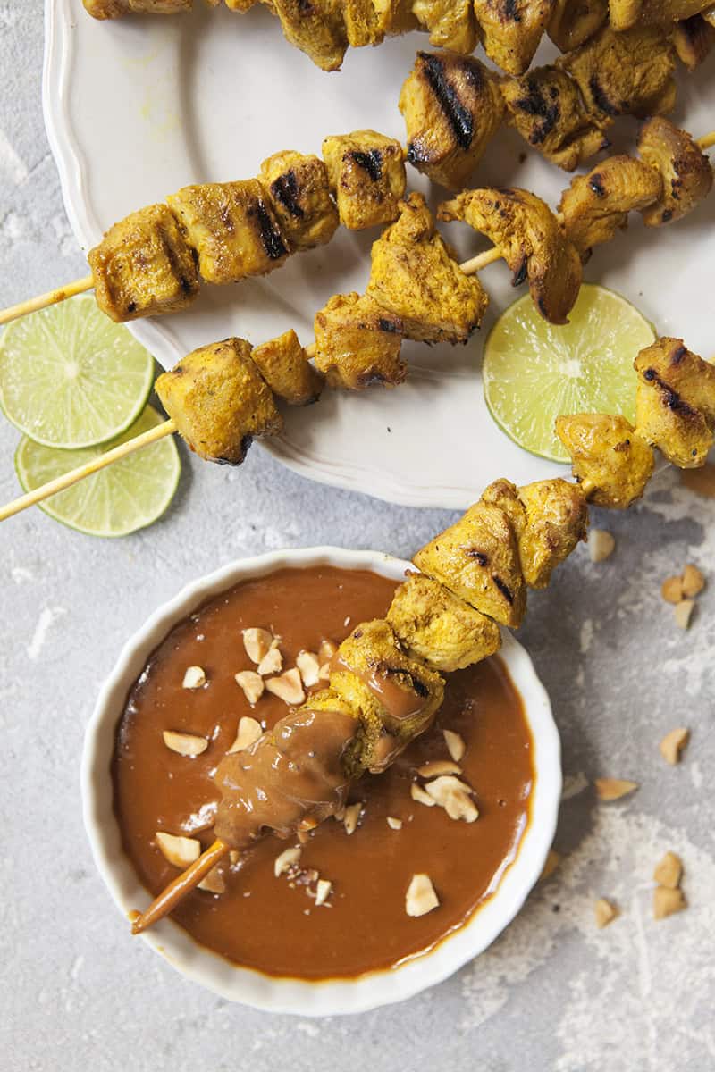 Thai-style grilled chicken satay skewers on a white scalloped plate alongside a bowl of peanut dipping sauce and chopped peanuts, slices of lime throughout the photo, on a gray background.