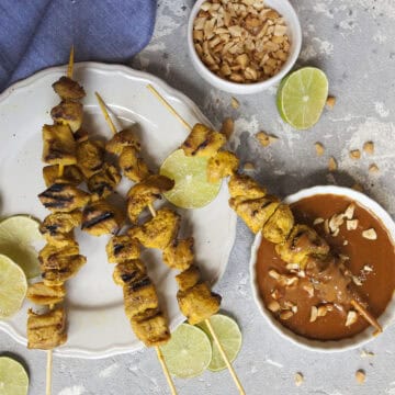 Thai-style chicken satay skewers on a white plate alongside a small bowl of peanut dipping sauce, lime slices, and a blue napkin on a gray background.