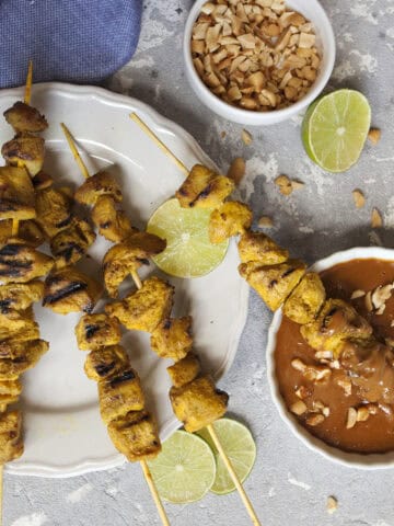 Thai-style chicken satay skewers on a white plate alongside a small bowl of peanut dipping sauce, lime slices, and a blue napkin on a gray background.