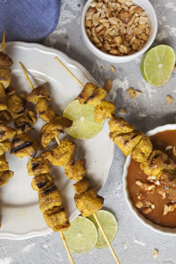 Thai-style chicken satay skewers on a white plate alongside a small bowl of peanut dipping sauce, lime slices, and a blue napkin on a gray background.