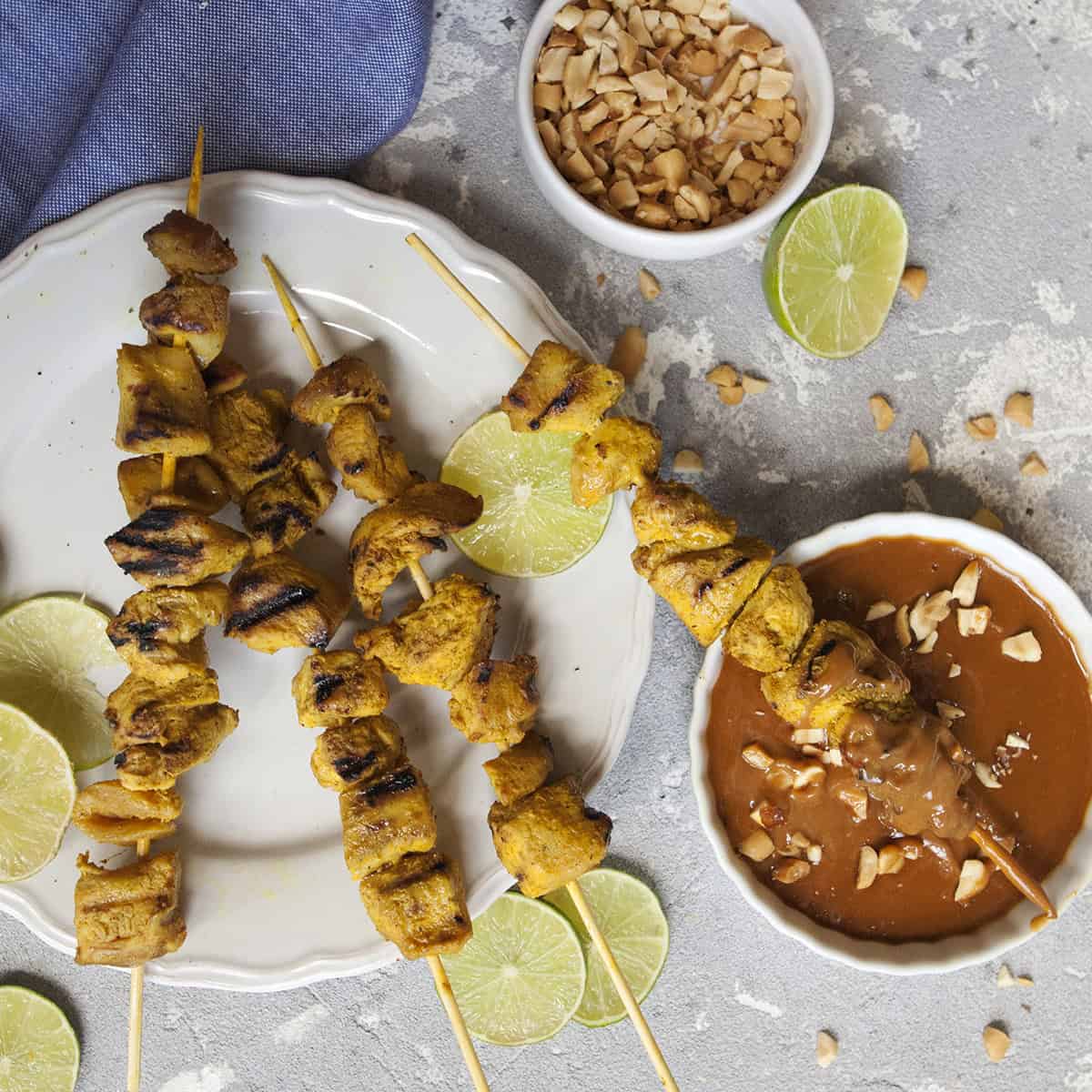 Thai-style grilled chicken satay skewers on a white scalloped plate alongside a bowl of peanut dipping sauce and chopped peanuts, slices of lime throughout the photo, on a gray background with a blue napkin.