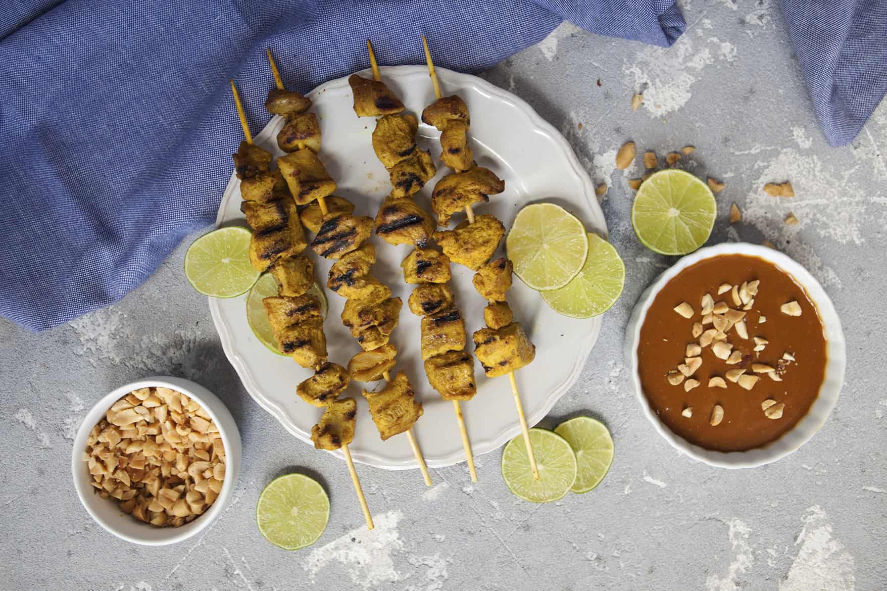 Thai-style grilled chicken satay skewers on a white scalloped plate alongside a bowl of peanut dipping sauce and chopped peanuts, slices of lime throughout the photo, on a gray background with a blue napkin.