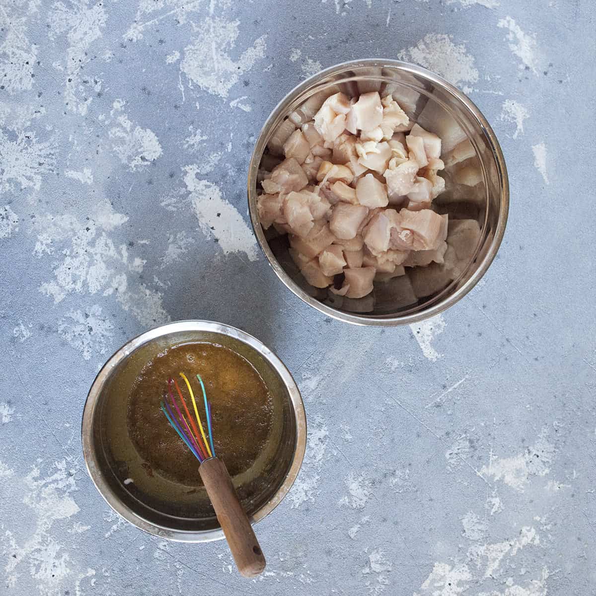 Chopped chicken breast in a bowl alongside a bowl of marinade ingredients and a whisk on a gray background.