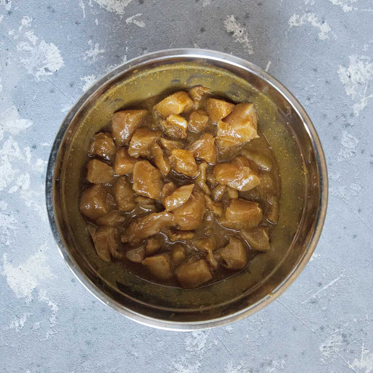 Chopped chicken pieces marinating in a brown sauce in a metal bowl on a gray background.