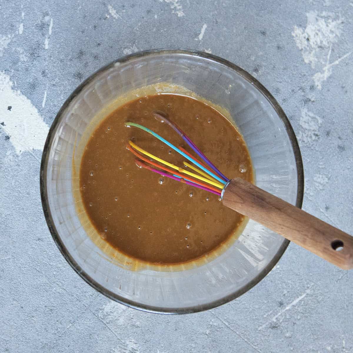 Medium brown, shiny peanut dipping sauce in a glass bowl with a whisk on a gray background.