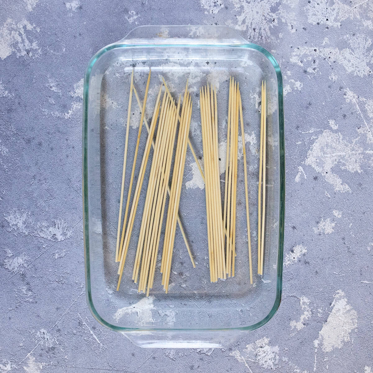 Wooden skewers soaking in water in a baking dish on a gray background.