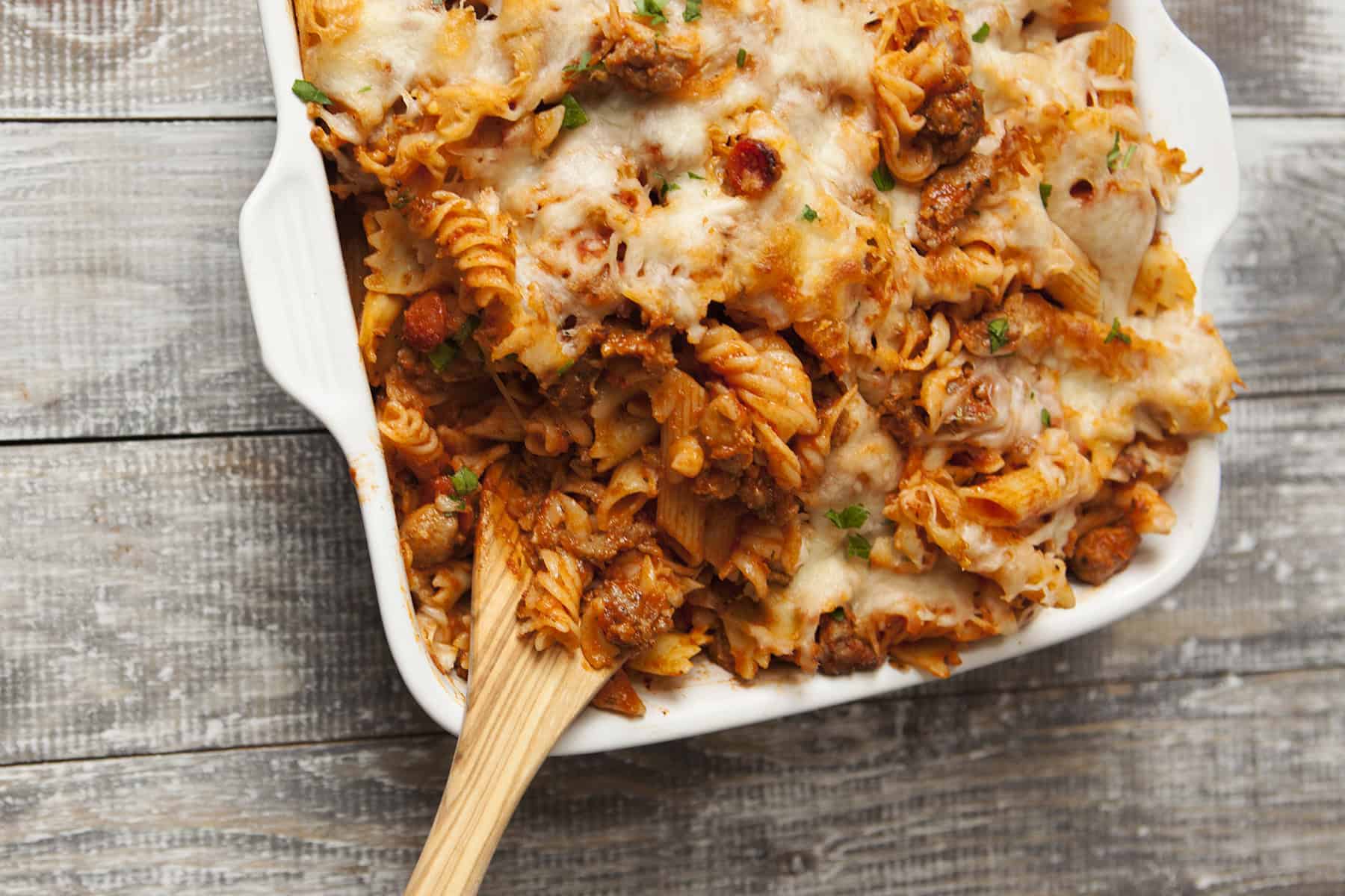 Pepperoni pasta bake with Itailan sausage and mozzarella in a white baking dish with a wooden spoon.