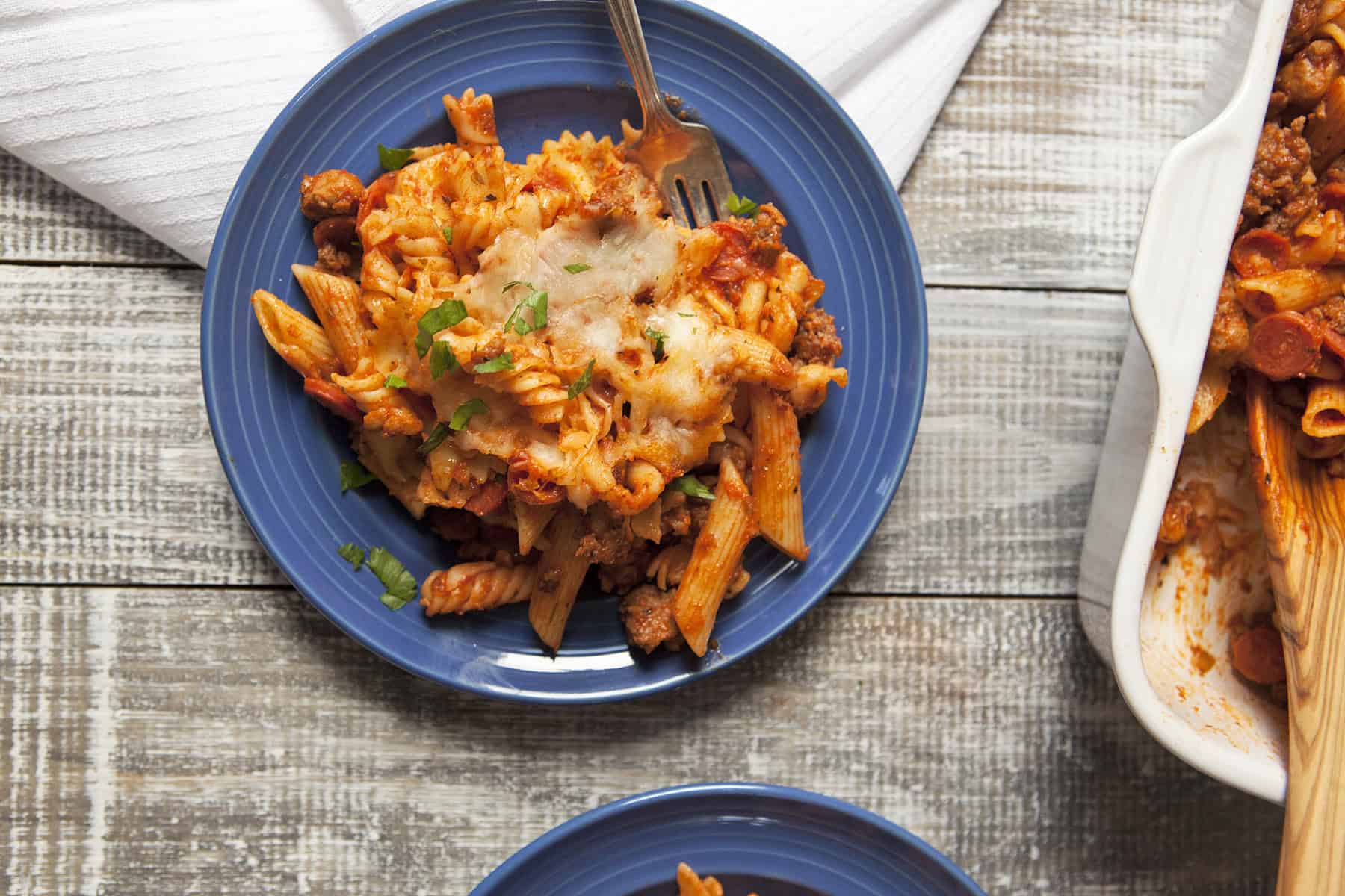 Pepperoni and Italian sausage pasta bake with red sauce on a blue plate, topped with chopped parsley, with a fork, on a table next to a white napkin and baking dish with more pasta bake.