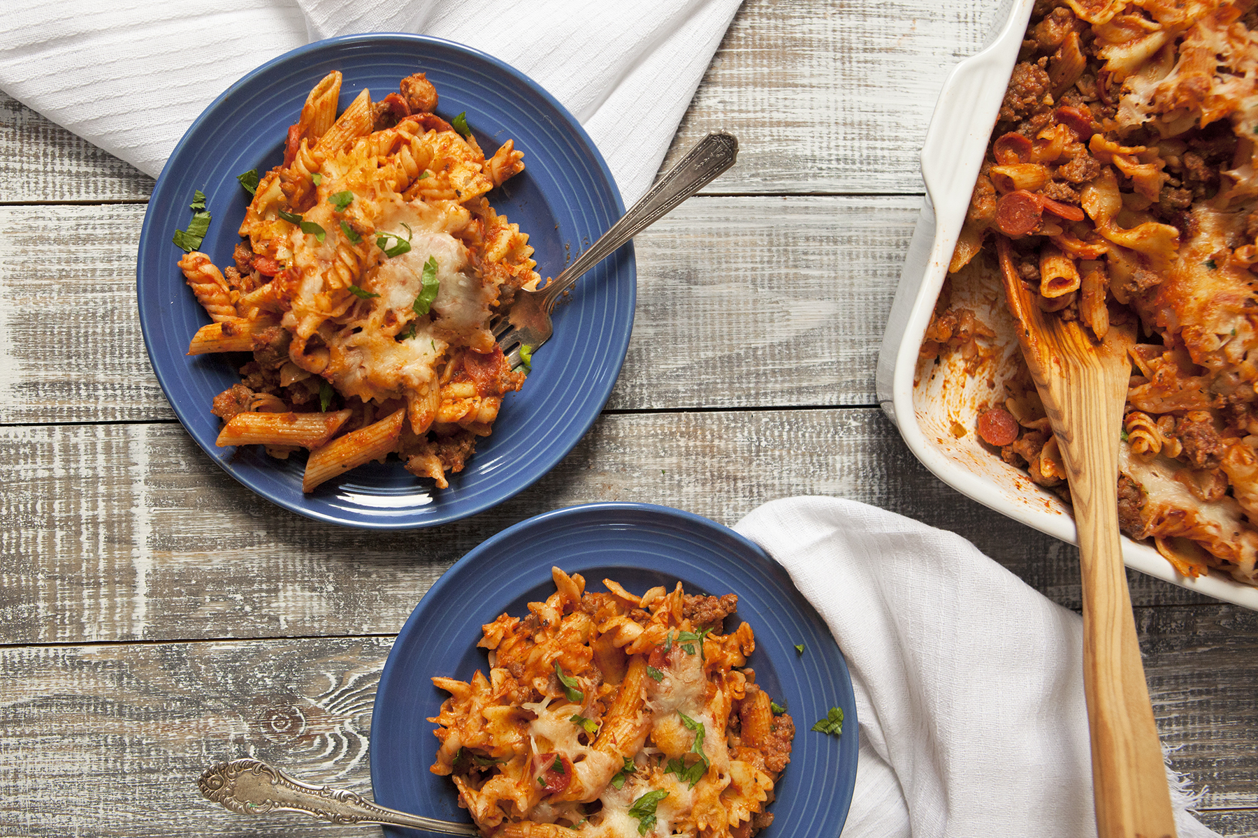 Two blue plates containing pepperoni pasta bake with Italian sausage and mozzarella with forks on a wooden table next to white fringed napkins and a white baking dish with more pepperoni pasta bake and a wooden spoon.