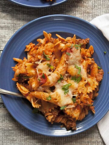 Pepperoni and Italian sausage pasta bake with red sauce on a blue plate next to a white napkin on a wooden table.