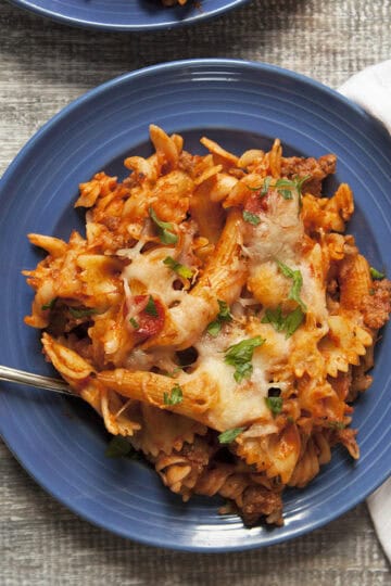 Pepperoni and Italian sausage pasta bake with red sauce on a blue plate next to a white napkin on a wooden table.