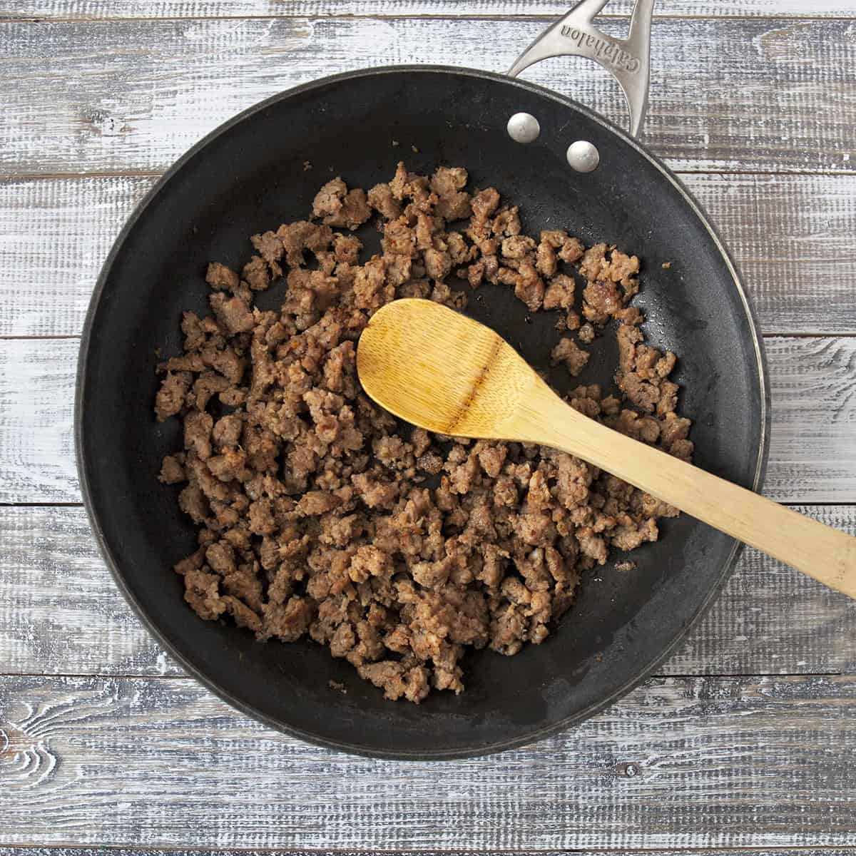 Ground Italian sausage browning in a skillet with a wooden spoon.