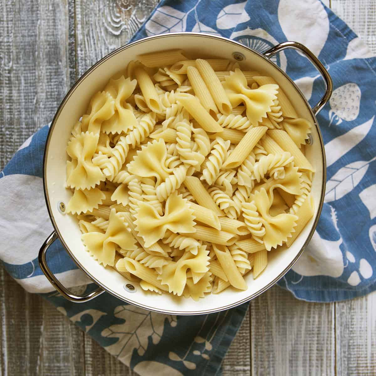 Pasta shapes (bowtie, rotini, and rigatoni) in a colander next to a blue and white kitchen towel.