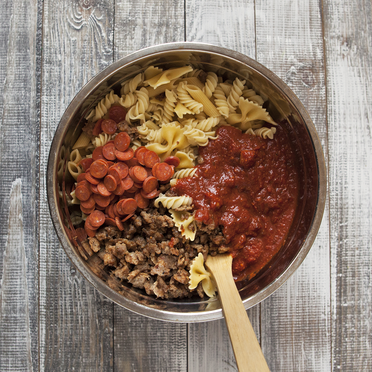 Ingredients for pepperoni pasta bake (cooked pasta, marinara sauce, mini pepperoni, and cooked ground Italian sausage) in a mixing bowl with a wooden spoon.