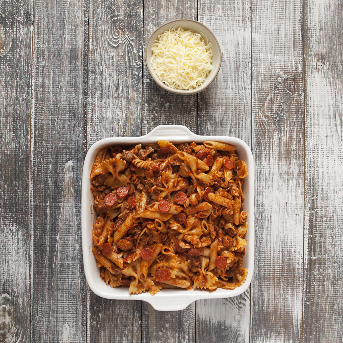 Pepperoni pasta bake in a square white baking dish next to a dish with grated mozzarella.