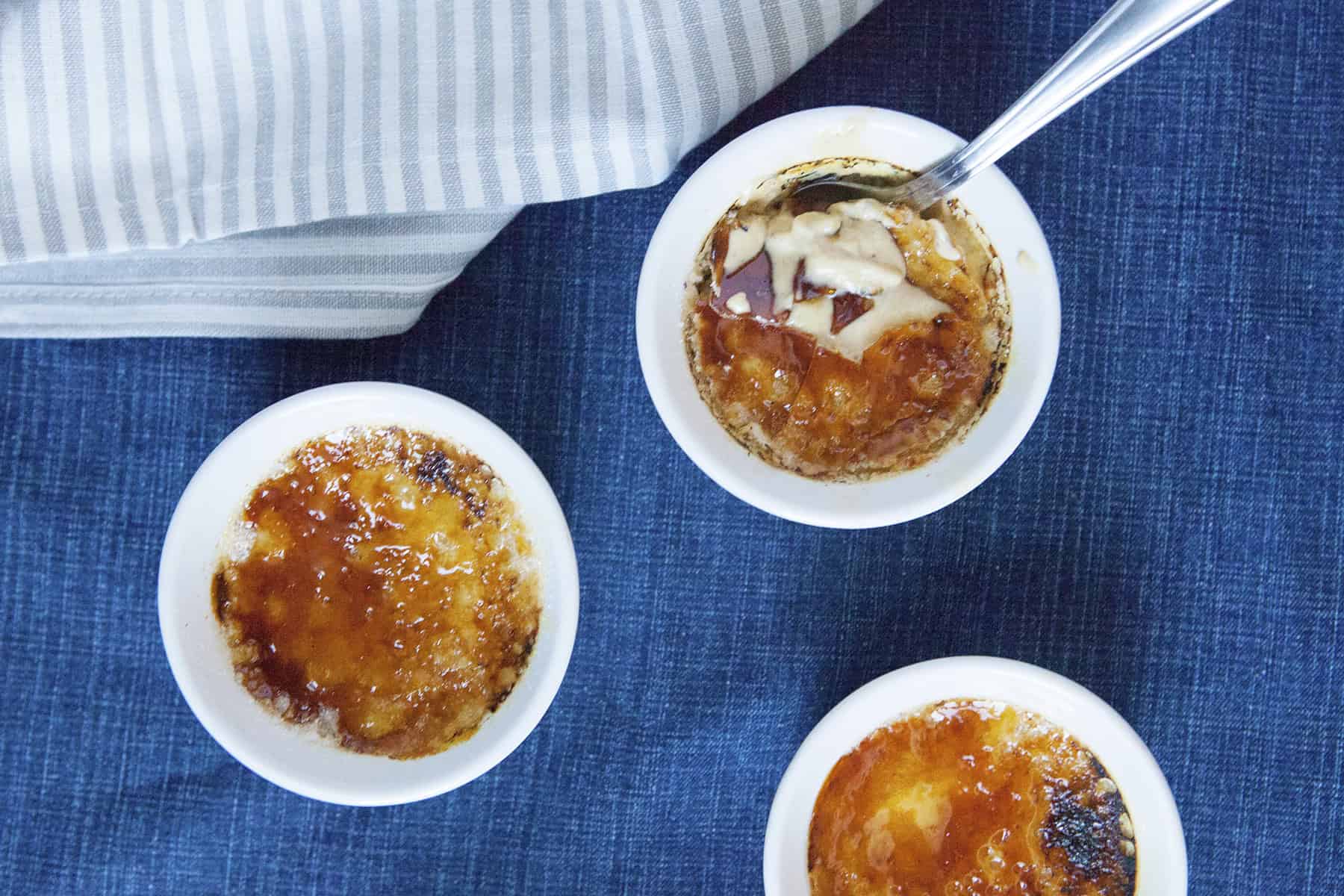 Chai-spiced creme brulee in 3 white ramekins with a spoon on a blue background with a gray napkin.