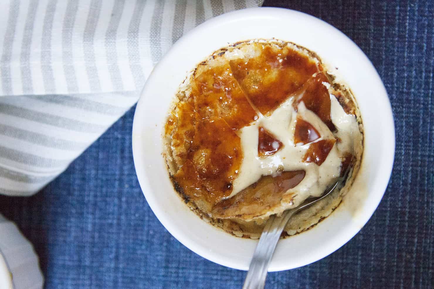 Chai-spiced creme brulee in a white ramekin with a spoon on a blue background with a gray napkin.