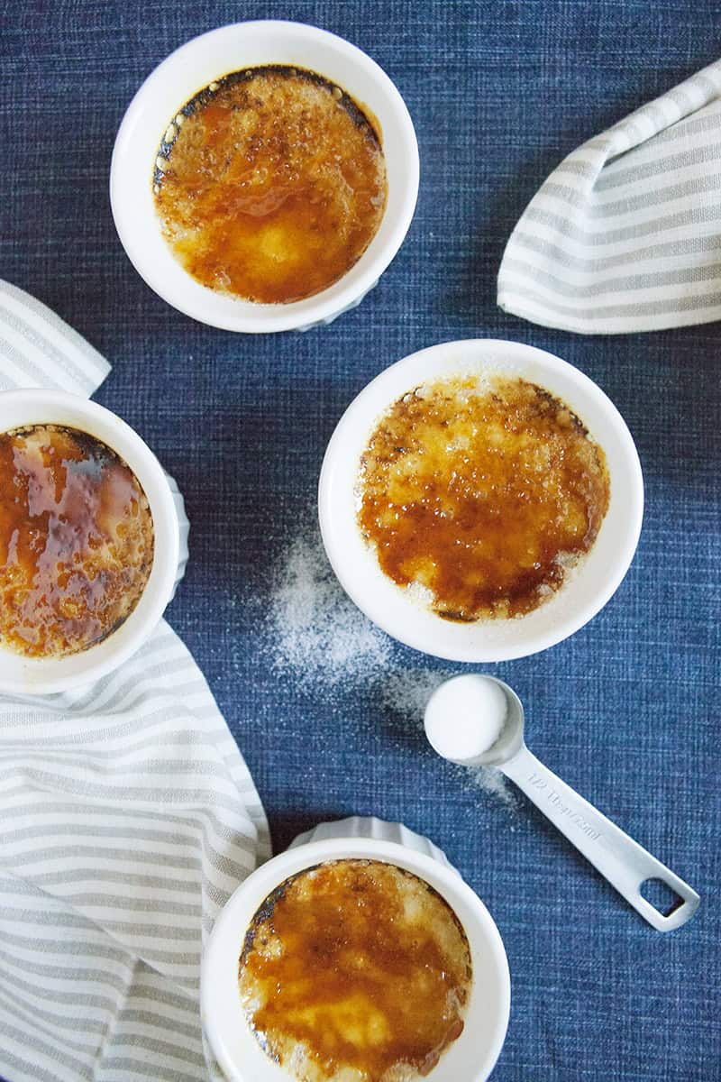 Chai-spiced creme brulee in 4 white ramekins with a measuring spoon containing granulated sugar on a blue background with a gray napkin.