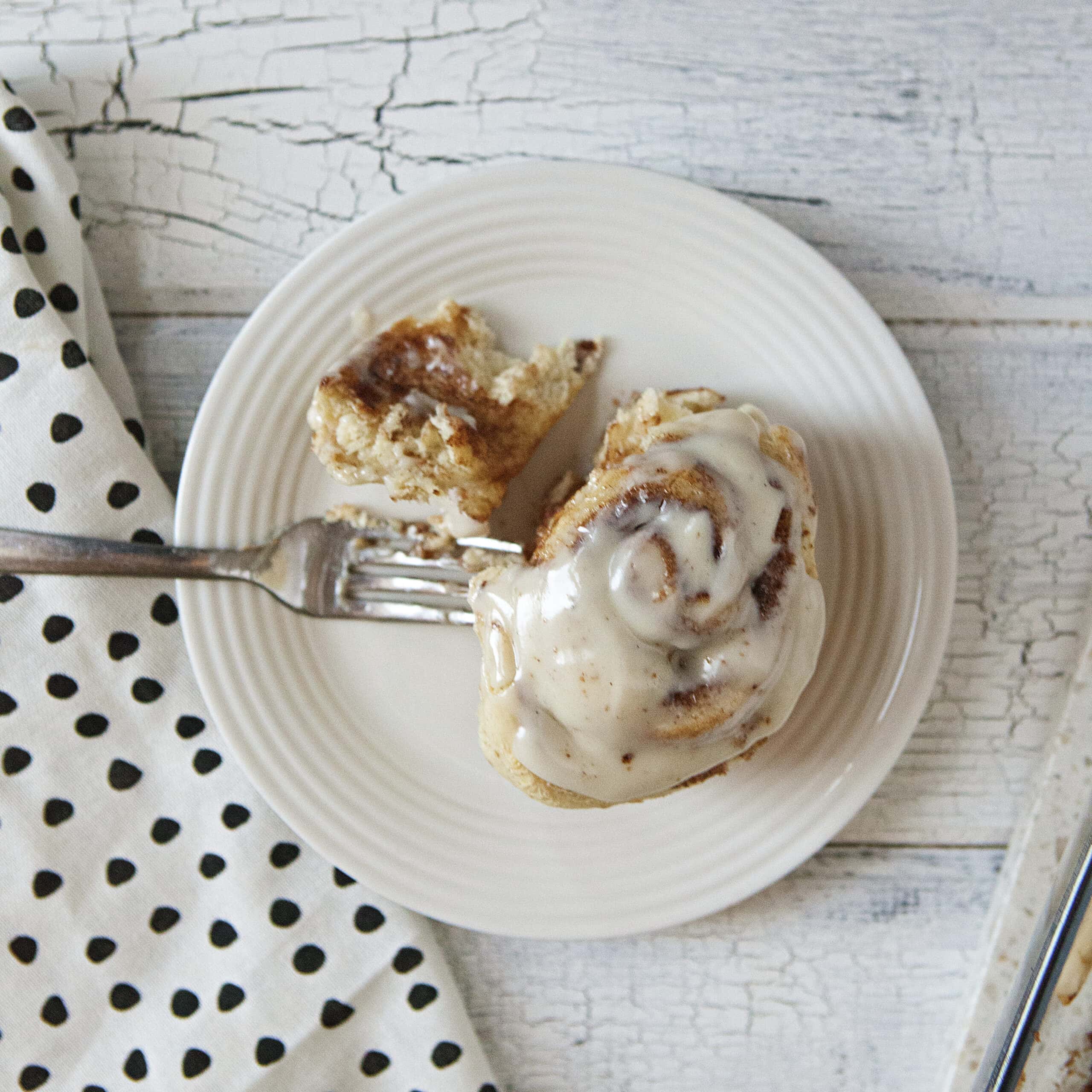 Soft and fluffy cinnamon roll with cream cheese frosting on a white plate, with a portion cut off with a knife, on a white wood background next to a white napkin with black polka dots.