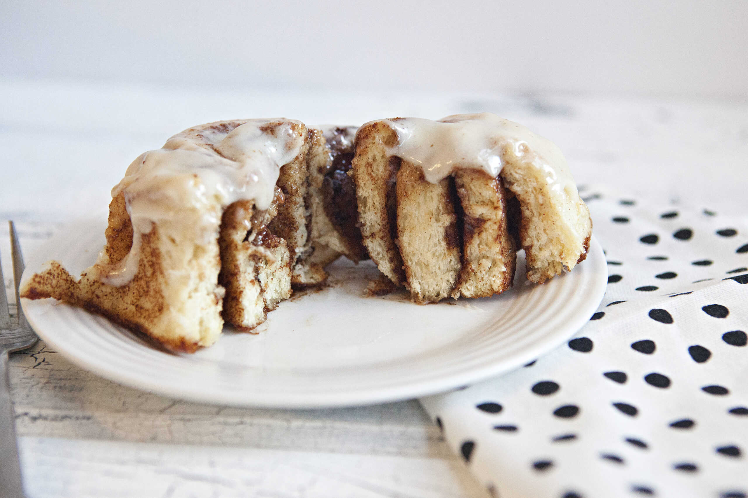 A soft and fluffy cinnamon roll with cream cheese frosting cut int half, showing the layers of dough and cinnamon filling, on a white plate next to a white napkin with black polka dots, alongside a fork.