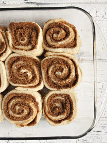 Soft and fluffy unbaked cinnamon rolls in a glass baking dish on a white wood background.