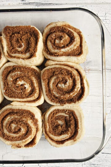 Soft and fluffy unbaked cinnamon rolls in a glass baking dish on a white wood background.